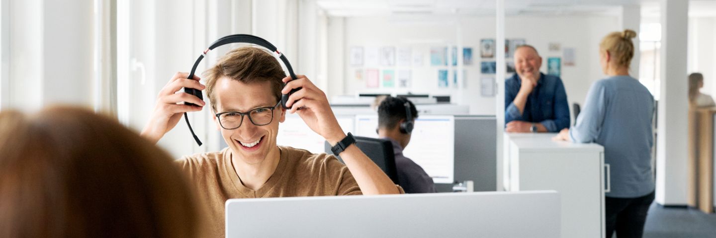 Un employé d'EOS avec des écouteurs travaille sur un ordinateur dans un bureau moderne, tandis que des collègues parlent en arrière-plan.
