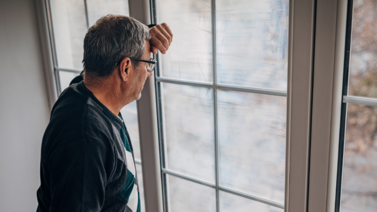 Recouvrement équitable des créances : un débiteur d'âge moyen vêtu d'une veste olive regarde par la fenêtre avec inquiétude.